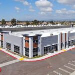 A modern industrial complex with multiple units, large windows, and ample parking. The building is surrounded by a clear, sunny sky with scattered clouds.