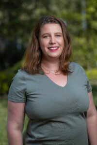 Erin Devlin, a woman with shoulder-length brown hair and wearing a gray t-shirt, is smiling outdoors with a blurred green background.