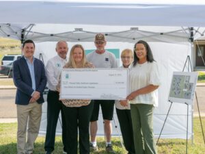 A group of people stand under a tent canopy, holding a large check for $3,150,000 from the U.S. Department of Housing and Urban Development, dated August 21, 2024. The funds are earmarked for improvements to Pleasant Valley Apartments.