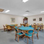 A room with several round tables, each surrounded by colorful chairs, evokes the charm of Sacramento Manor. Framed pictures and a cabinet with decor adorn the walls. The space is well-lit with overhead lighting, adding to its inviting atmosphere.