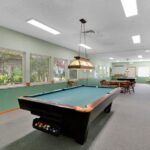 The recreational room at Sacramento Manor features two pool tables, a chalkboard, chairs, windows, and framed pictures on green walls under fluorescent lights.