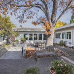 A courtyard at Sacramento Manor features a large tree shedding autumn leaves, surrounded by a single-story building, benches, plants, and a pathway with a ramp.