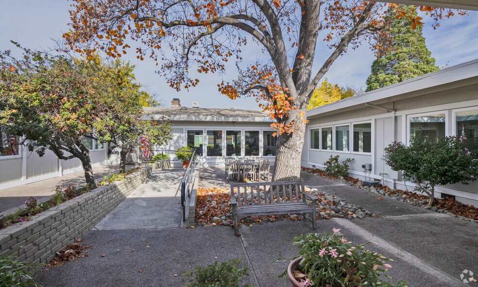 A courtyard at Sacramento Manor features a large tree shedding autumn leaves, surrounded by a single-story building, benches, plants, and a pathway with a ramp.