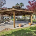 A wooden gazebo with benches inside is situated in the picturesque Sacramento Manor park. There are potted plants in the gazebo and trees nearby with vibrant autumn foliage. A path winds through the surrounding green lawn, creating a serene and inviting atmosphere.