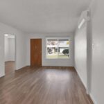 A vacant room at Sacramento Manor features white walls, wood flooring, a wooden front door, a window, and a small air conditioning unit on the wall.