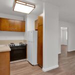 A compact kitchen at Sacramento Manor features wooden cabinets, white countertops, a white refrigerator, and a small oven. The adjacent living area boasts wood flooring and a charming wooden front door.