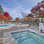 The outdoor pool area at Sacramento Manor features lounge chairs, a hot tub, and an accessibility lift. Surrounded by trees with red and orange leaves, it offers a serene escape with residential buildings in the background.