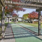 Green shuffleboard courts under pergolas in a park setting at Sacramento Manor, with trees and buildings in the background on a sunny day.