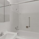 A clean white bathroom at Sacramento Manor features a bathtub with white tiled walls, a sink with a chrome faucet, and grab bars for assistance.